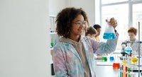 Young African American female scientist examining blue chemical solution in laboratory with colleagues conducting research experiments in background.
