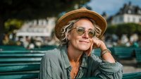 Relaxed senior woman with gray hair wearing straw hat and sunglasses sitting on park bench, smiling peacefully in urban setting for lifestyle content.
