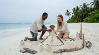 Young mixed-race family building sandcastle together on tropical beach with palm trees and turquoise water, parents and child enjoying summer vacation and outdoor recreation activities.