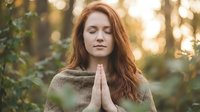 Young Caucasian woman with long red hair meditating outdoors in prayer pose with eyes closed, peaceful expression in natural forest setting at golden hour.