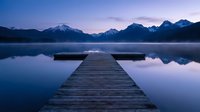 Wooden dock extending into calm mountain lake at twilight with snow-capped peaks reflecting in still water creating peaceful alpine scenery.