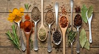 Colorful spices and herbs arranged in wooden spoons on rustic wood background. Cooking ingredients including turmeric, paprika, coriander, peppercorns, bay leaves.