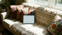 Tablet with blank white screen on a boho-style sofa with colorful pillows and macrame blanket in a sunny living room.
