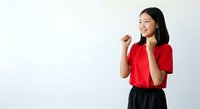 Young Asian woman wearing bright red shirt and black skirt, smiling with hands raised in celebratory gesture against white background, expressing joy and success.