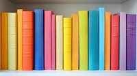 Row of colorful hardcover books standing on wooden shelf against gray wall background, spines showing rainbow arrangement from yellow through blue to purple.