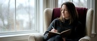 Young woman reading book in cozy armchair by rainy window. Peaceful indoor leisure activity on gloomy day. Relaxation and literature concept.