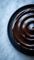 Rich chocolate ganache swirl in dark bowl on marble surface for baking and dessert preparation.