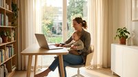 Young Caucasian woman working from home office with laptop computer sitting at wooden desk near window with natural sunlight and bookshelves background.