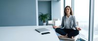 Young professional businesswoman meditating in lotus position on modern office desk during work break for stress relief and mindfulness practice.