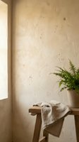 Minimalist interior with cream textured wall, natural light from window, potted fern plant, and folded linen cloth on wooden stool for home decor and lifestyle design projects.