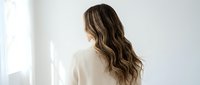 Young woman with long wavy brown hair wearing white shirt standing by window in bright minimalist room for wellness and lifestyle concepts.