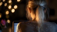 Young woman working late at computer with glowing bokeh lights in background, wearing sparkly sweater, blonde hair in messy bun, creating cozy evening atmosphere.