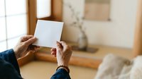 Person with dark skin tone holding blank white card in minimalist interior space with natural light, suitable for greeting cards, invitations, announcements, and marketing mockups.