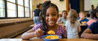 Young African American girl with braids smiling while eating lunch in school cafeteria with diverse classmates in background during meal time.