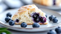 Fresh blueberry scone with glazed top served on white plate, garnished with fresh berries. Close-up view shows moist, flaky texture and juicy fruit filling.