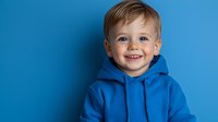 Cheerful toddler boy wearing bright blue hoodie smiling against blue background. Close up portrait showing genuine happiness and positive emotions.