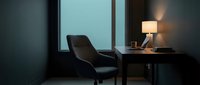 Modern dark interior with comfortable armchair positioned by large window at dusk, minimalist desk with lamp, contemporary home office or bedroom design concept.