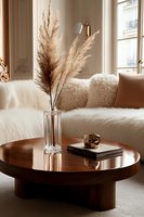 Cozy living room corner with pampas grass in crystal vase on round wooden coffee table, white sheepskin throws and cushions create warm minimalist atmosphere.