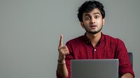 Young South Asian male professional in red checkered shirt gesturing while working on laptop against gray background, expressing surprise and insight during remote work.