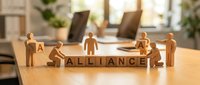 Wooden figures arranged around letter blocks spelling alliance on office desk with laptops and plants in background, representing business partnership and teamwork.