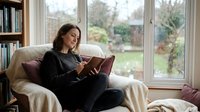 Young woman reading book on comfortable sofa by large window with garden view. Cozy home interior with natural light for relaxation and leisure lifestyle.
