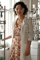 Young Asian female scientist in laboratory wearing protective glasses and white coat over floral dress, standing next to chemical equipment and glassware. Professional research setting.