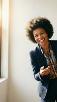 Young African American woman with natural curly hair wearing navy blazer and patterned scarf, smiling warmly while gesturing welcomingly near bright window for business and corporate use.