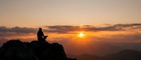 Person silhouette meditating on mountain peak during golden sunset with dramatic sky and layered mountain ranges creating peaceful spiritual atmosphere.