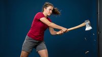 Young Caucasian woman in red shirt swinging axe with determination and focus. Action shot shows strength, power and outdoor adventure activity.