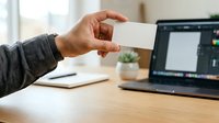 Hand holding blank white business card at wooden desk with laptop and notebook, suitable for branding and corporate identity presentations.