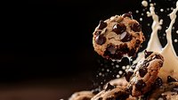 Fresh baked chocolate chip cookie breaking apart with milk splash and droplets against dark background, dramatic food photography composition.