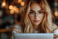 Young blonde woman with glasses working on laptop in cozy cafe with warm bokeh lights. Professional freelancer or student studying in comfortable workspace.