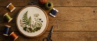 Embroidery hoop with botanical fern and wildflower design surrounded by colorful thread spools and scissors on rustic wooden table for crafting projects.