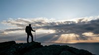 Silhouette of hiker with trekking poles standing on rocky mountain peak at sunset, dramatic cloudy sky with golden sunrays, outdoor adventure and exploration concept.