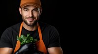 Smiling male chef in orange apron and black t-shirt holding fresh herbs against dark background, professional culinary portrait with dramatic lighting.