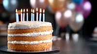 Layered vanilla birthday cake with lit blue and white candles on dark background, colorful party balloons bokeh. Celebration dessert for special occasion.