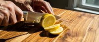 Hands slicing fresh lemon with knife on wooden cutting board in bright sunlight, food preparation for cooking and beverage recipes.