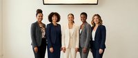 Diverse group of five professional businesspeople standing together in modern office setting wearing formal blazers and business attire.