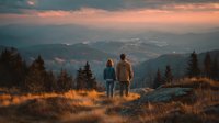 Couple hiking at mountain summit during golden hour sunset with panoramic valley view and dramatic sky creating romantic outdoor adventure moment.