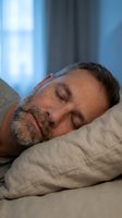 Middle-aged man with beard sleeping peacefully on pillow with eyes closed, resting in bedroom with natural light from window for sleep and wellness content.