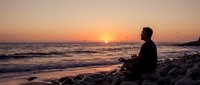Man meditating in lotus position on rocky beach at sunset with ocean waves and flying birds creating peaceful mindfulness scene for wellness content.