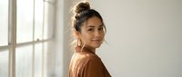 Young Asian woman with hair bun wearing brown sweater smiling at camera in bright modern interior with natural window light for lifestyle content.