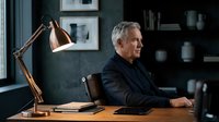 Mature man with gray hair, Caucasian ethnicity, wearing dark blazer, seated at wooden desk in modern office with brass lamp, tablet, and dark shelving. Professional workspace setting.