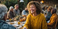 Middle-aged woman with curly hair laughing during outdoor family dinner gathering, wearing yellow knit sweater at festive autumn table.