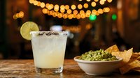 Refreshing margarita cocktail with lime slice and salt rim served with guacamole dip and tortilla chips at festive bar setting with warm string lights.