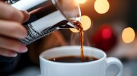 Fresh coffee pouring from metal thermos into white ceramic cup with warm bokeh lights in background, creating cozy morning atmosphere.