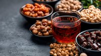Glass of traditional red tea surrounded by various dried fruits and nuts in black ceramic bowls on dark stone background, middle eastern style snacks and drinks.