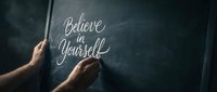 Hands writing motivational quote believe in yourself on dark chalkboard with chalk for inspiration and self-confidence concepts.