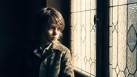 Young boy with tousled hair standing by ornate stained glass window in warm sunlight, creating dramatic shadows and peaceful contemplative mood.