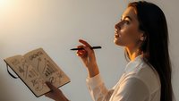 Young Hispanic businesswoman in white shirt holding notebook with creative diagrams, thinking with pen in hand during brainstorming session.